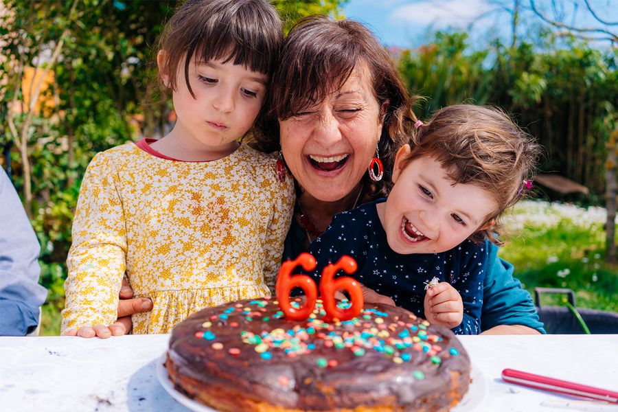 Une femme sourit près de son gâteau d'anniversaire orné de bougies indiquant « 66 », entourée de deux enfants souriants, illustrant les conseils de Photobox pour capturer les moments d'anniversaire intergénérationnels.