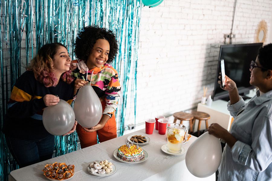 Deux filles posent devant un fond de photo en tinsel, tenant des ballons près d'une table de snacks garnie de nourriture de fête et d'un gâteau d'anniversaire, illustrant les conseils de Photobox pour photographier la décoration et les buffets de fête.