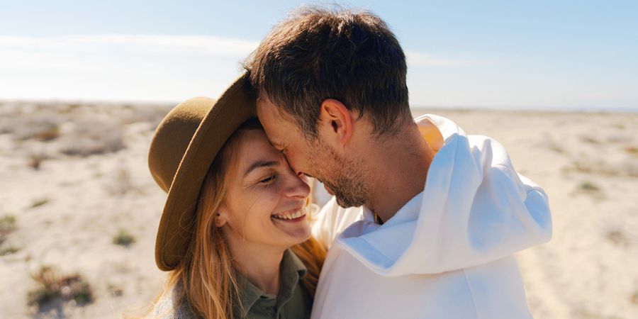 Un couple dans la trentaine sourit et s'enlace dans un décor de plage désertique, présentant le guide de Photobox de 60 idées de messages pour une carte d'anniversaire de mariage.