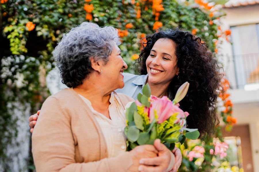 Une jeune femme sourit à sa mère qui tient des fleurs, illustrant les idées de Photobox pour savoir quoi écrire dans une carte d'anniversaire de mariage pour ses parents.