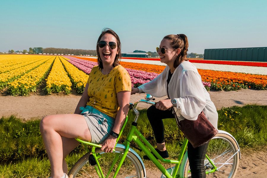Deux femmes font du vélo ensemble, souriant et riant dans un champ rempli de fleurs, illustrant les idées de Photobox pour savoir quoi écrire dans une carte d'anniversaire de mariage pour des amis.
