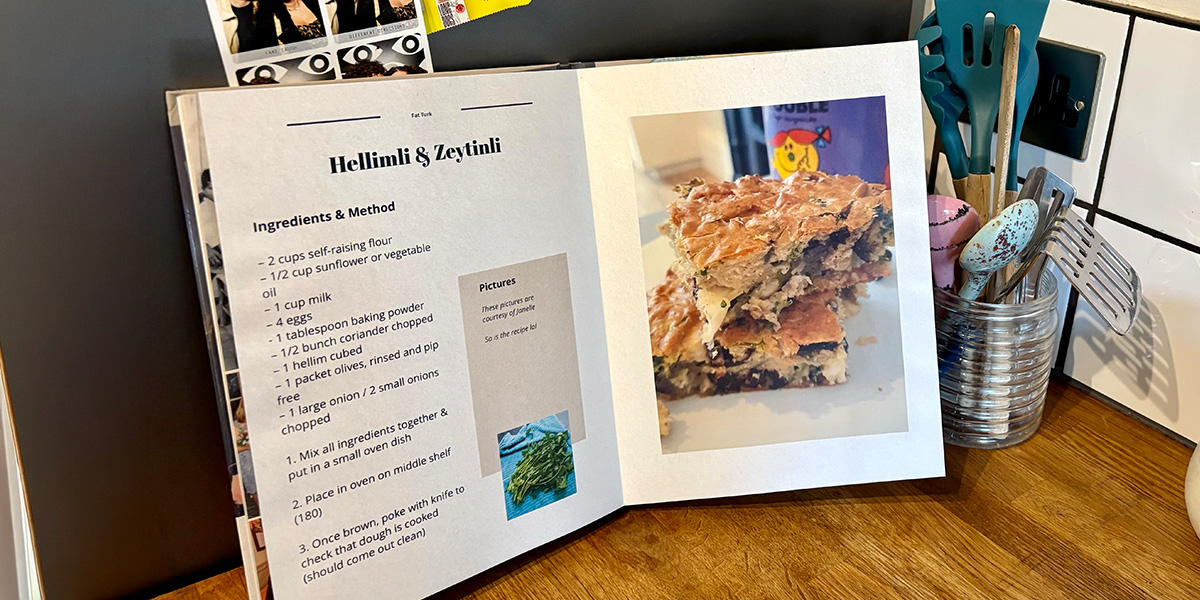 Un livre photo recette ouvert est posé dans une cuisine, avec un pot de cuillères et d'ustensiles de cuisine en arrière-plan, présentant le guide de Photobox pour créer son propre livre de recettes.