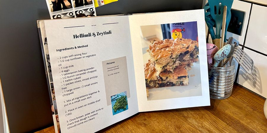 Un livre photo recette ouvert est posé dans une cuisine, avec un pot de cuillères et d'ustensiles de cuisine en arrière-plan, présentant le guide de Photobox pour créer son propre livre de recettes.