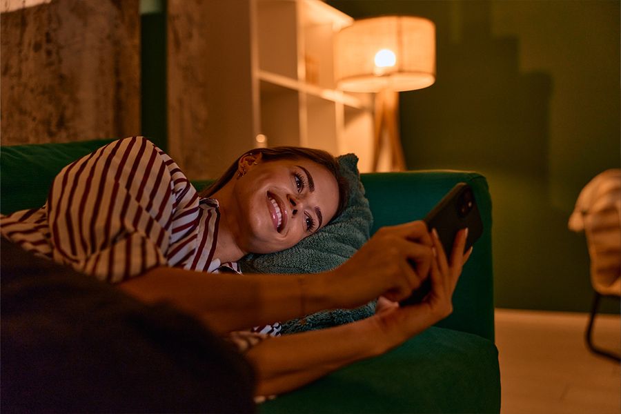 Une femme est allongée sur un canapé vert foncé et regarde son téléphone dans un salon, éclairée par une lampe pour une lumière chaleureuse, illustrant les conseils de Photobox pour un éclairage hygge à la maison.