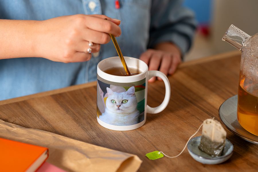 Quelqu'un remue une cuillère dans un mug photo personnalisé orné d'une photo de chat blanc en préparant une tasse de thé, illustrant le conseil de Photobox de compléter un coin lecture hygge avec un mug personnalisé.