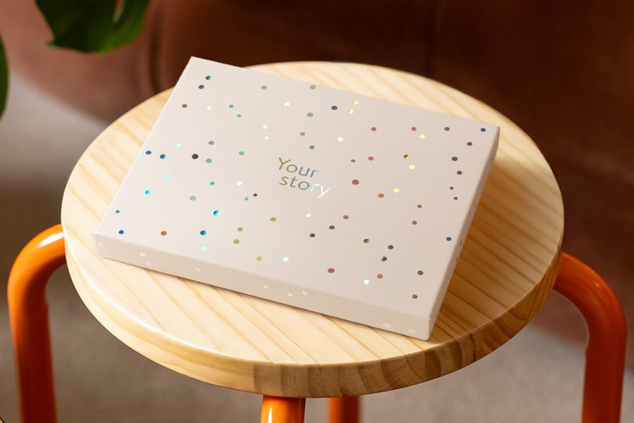 Une boîte de présentation Photobox est posée sur un tabouret en bois, décorée de cercles holographiques argentés et du texte « Your Story », illustrant le design mat ivoire avec une touche de brillance holographique.