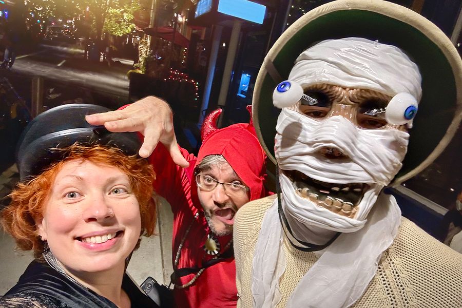 Trois adultes déguisés pour Halloween dans la rue, l'un avec un masque de squelette aux yeux exorbités, un autre en diable et une femme coiffée d'un chapeau noir, illustrant les idées de légendes et de phrases d'Halloween pour les déguisements de Photobox.