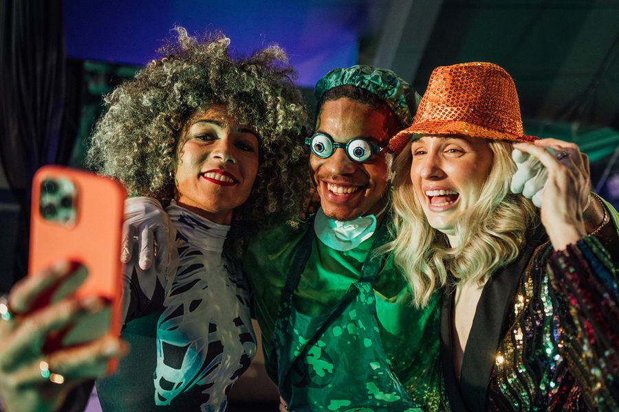 Trois adultes posent pour un selfie lors d'une soirée d'Halloween, une femme déguisée en squelette, un homme en chirurgien effrayant avec des lunettes rigolotes et une femme coiffée d'un chapeau à paillettes, illustrant les légendes et phrases d'Halloween de Photobox pour les groupes.