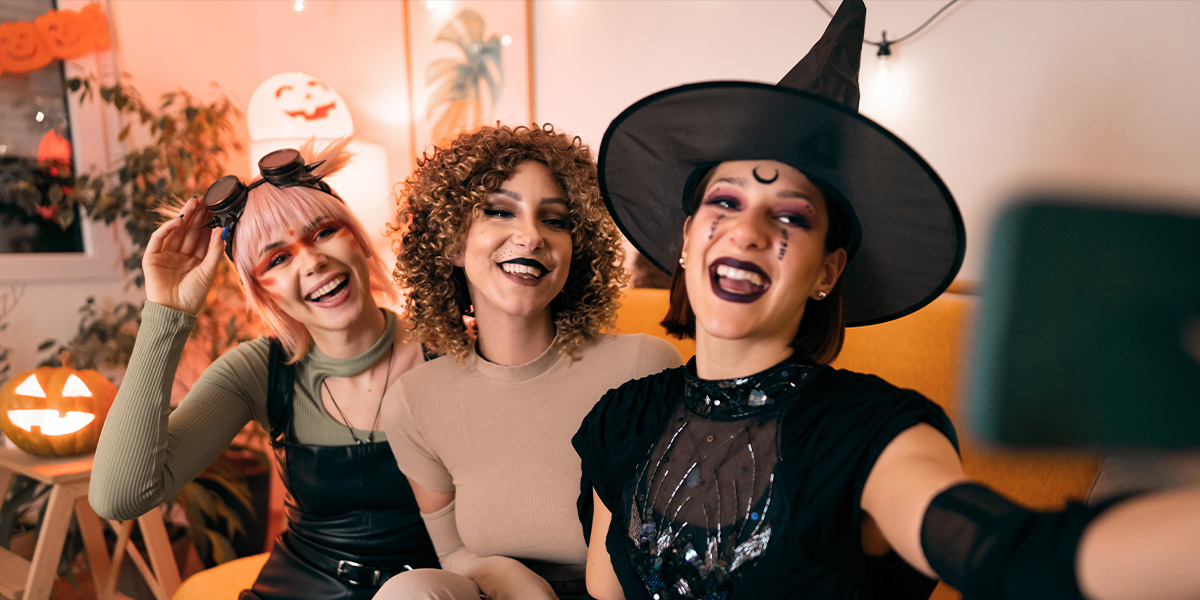 Trois femmes déguisées lors d'une soirée d'Halloween, une sorcière, un animal et une cyberpunk, posent pour un selfie sur leur téléphone, présentant les 105 citations et phrases d'Halloween pour Instagram de Photobox.