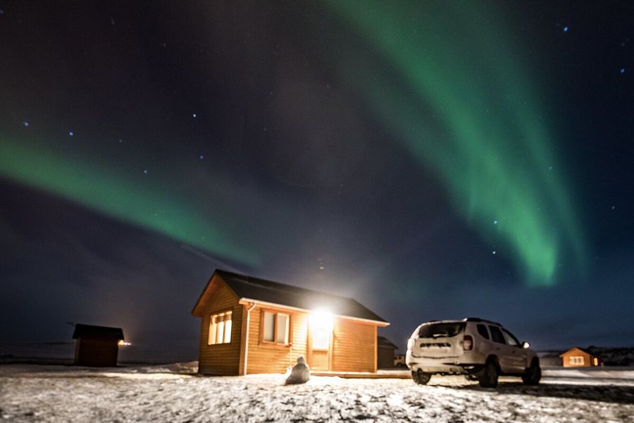 Une cabane avec une lumière qui brille à côté d’une voiture, dans un paysage très enneigé, avec des aurores boréales vertes dans le ciel au-dessus, illustrant l’explication de Photobox sur ce que sont les aurores boréales et ce qui les provoque.