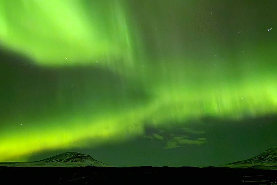 Montagnes enneigées sous un ciel d’un vert intense grâce aux aurores boréales, illustrant les conseils de Photobox pour voir les aurores boréales ailleurs qu’en Islande.