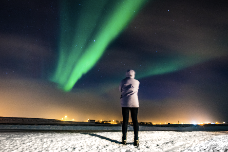 Une personne debout dans un paysage enneigé, face aux aurores boréales vertes dans le ciel, illustrant les conseils de Photobox pour photographier une aurore boréale avec un smartphone.