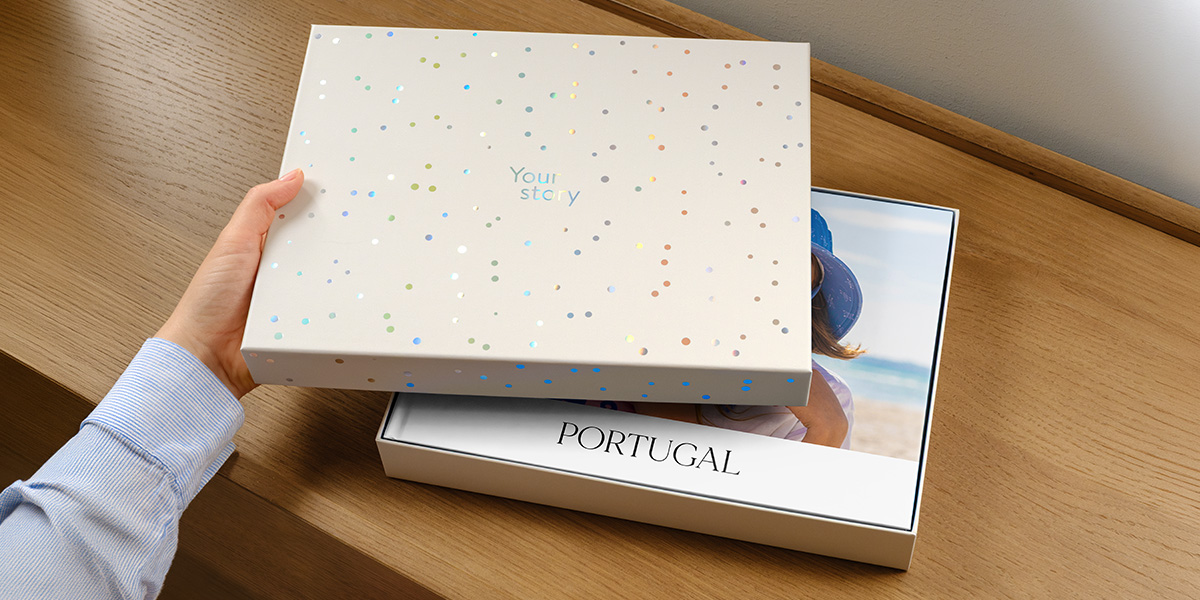 Une boîte de présentation Photobox est posée avec son couvercle légèrement entrouvert, révélant un livre photo de voyage personnalisé pour le Portugal à l'intérieur, dont la couverture est partiellement cachée par le couvercle, présentant la nouvelle boîte cadeau pour livre photo de Photobox.