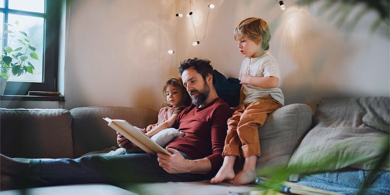 Un père lit un livre à un jeune garçon et à une petite fille sur le canapé, avec des guirlandes lumineuses chaudes devant eux, présentant les conseils de Photobox pour une déco d'automne hygge et chaleureuse.