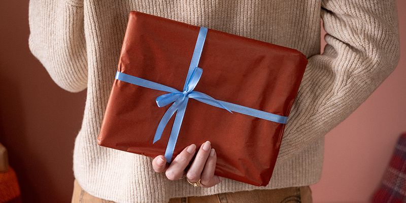 Femme tenant derrière son dos un cadeau emballé dans du papier marron avec un ruban bleu, pour illustrer la checklist de Photobox pour trouver le cadeau parfait de Noël avec un cadeau photo personnalisé.