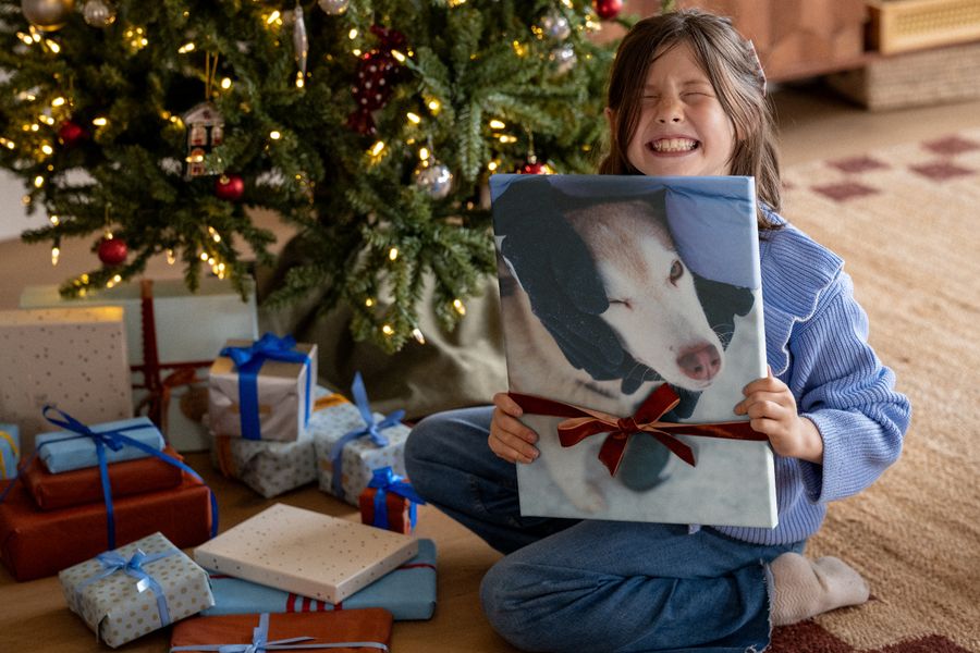 Une fillette riante en pull bleu tient un tirage sur toile d’un chien, noué d’un ruban rouge, devant un sapin de Noël, illustrant les idées de décoration murale de Photobox pour les photos qui méritent une place d’honneur.