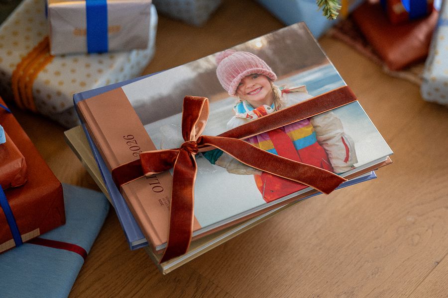 Pile de livres photo personnalisés entourés d’un ruban en velours marron, avec d’autres cadeaux emballés en arrière-plan, pour illustrer les conseils de Photobox pour préparer un cadeau photo personnalisé à Noël.