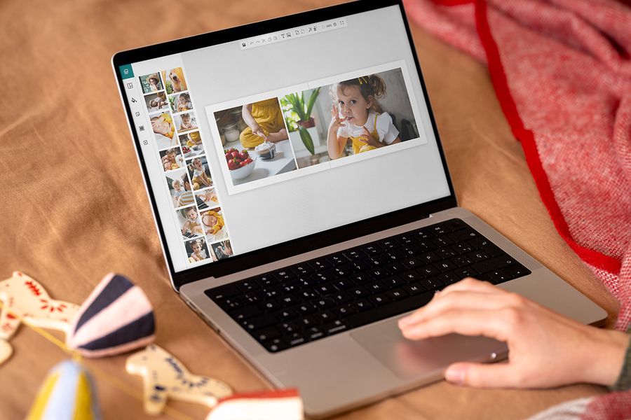 Une main sur le pavé tactile d’un ordinateur portable crée un livre photo à l’écran, avec une double page montrant une petite fille et un bol de fraises, illustrant l’éditeur en ligne de Photobox pour préparer des cadeaux de Noël pendant le Black Friday.