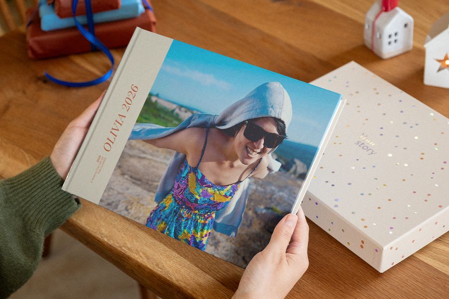 Deux mains tiennent un livre photo à couverture 2026 représentant une femme souriante en lunettes de soleil sur une côte rocheuse, à côté d’une boîte de présentation à motifs de confettis, illustrant les livres photo personnalisés de Photobox.