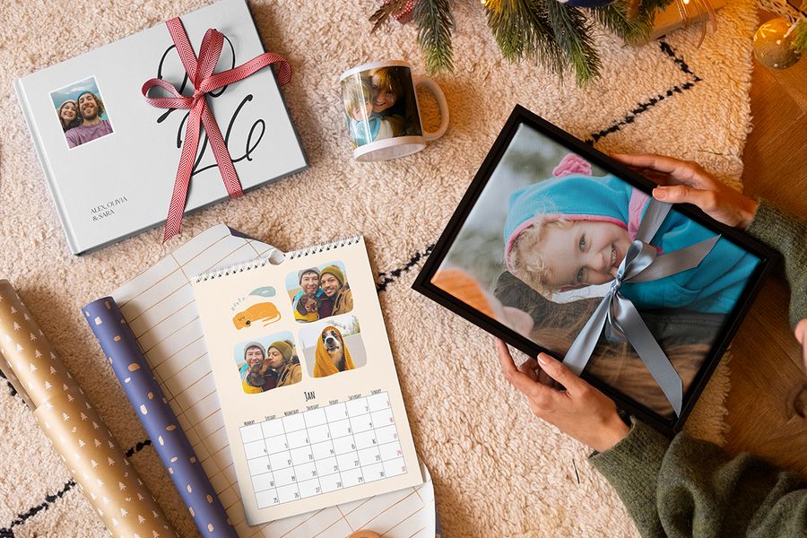 Sélection de cadeaux personnalisés Photobox, avec une décoration murale représentant une petite fille entourée d’un ruban bleu, un mug avec une mère et son enfant, un calendrier photo personnalisé et un livre photo 2026 entouré d’un ruban rouge, à côté de papier cadeau et avec des branches de sapin de Noël en arrière-plan, pour illustrer les conseils de Photobox pour capturer les bons souvenirs dans un cadeau de Noël personnalisé.