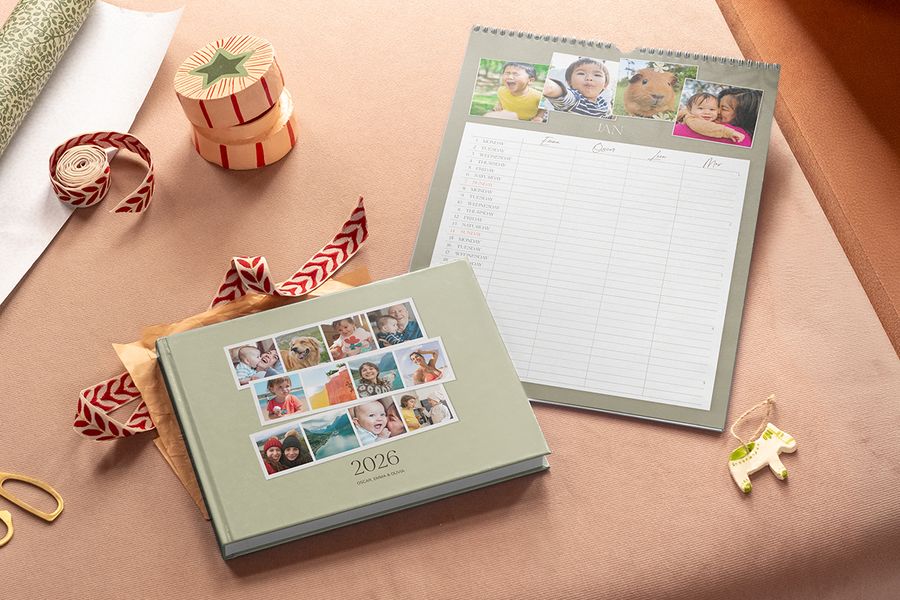 Un livre photo vert sauge à couverture 2026 est posé devant un calendrier familial mural orné de photos en haut, avec des colonnes pour chaque membre de la famille, illustrant le calendrier familial partagé de Photobox pour les cadeaux de Noël du Black Friday.