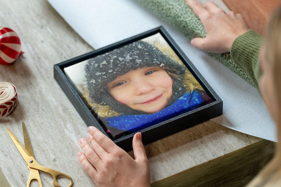Toile encadrée représentant une petite fille en vêtements d’hiver dans la neige, avec les mains d’une femme prêtes à l’emballer, à côté de papier cadeau, de ciseaux et de ruban, pour illustrer les conseils de Photobox pour choisir un cadeau photo personnalisé qui correspond au style de la personne.