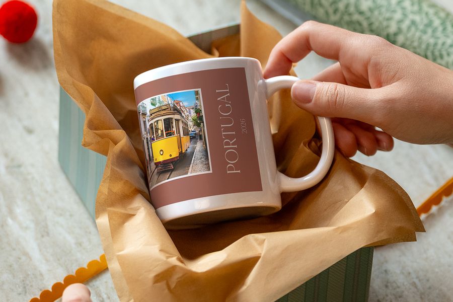 Une main soulève un mug emballé dans du papier kraft, imprimé d’un tram jaune et du titre « Portugal 2026 », illustrant les mugs personnalisés de Photobox comme indice pour un cadeau expérience.