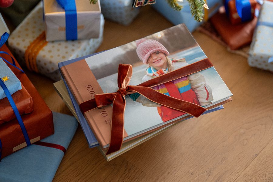 Une pile de livres photo nouée d’un ruban de velours, le livre du dessus montrant une fillette souriante en bonnet rose et « 2026 » sur la tranche, parmi des cadeaux de Noël emballés, illustrant le livre photo de l’année de Photobox.