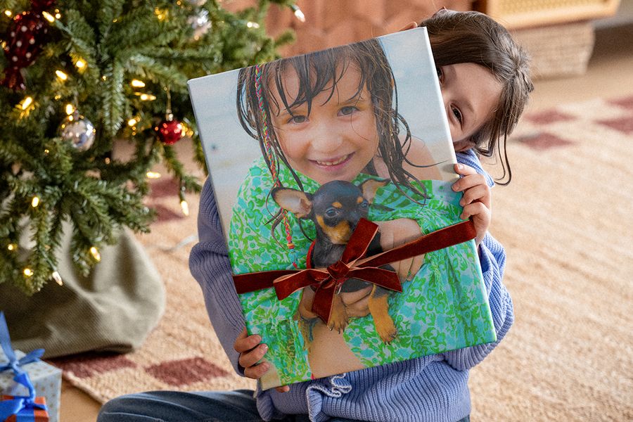 Petite fille sous le sapin de Noël tenant un grand livre photo entouré d’un ruban en velours, avec une photo d’elle et d’un chihuahua en couverture, pour illustrer la checklist de Photobox pour un cadeau photo personnalisé à Noël.