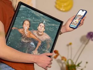 Une femme tient son téléphone affichant l’éditeur Photobox tout en portant sous son bras une décoration murale de vacances montrant deux hommes s’amusant dans une piscine, pour inspirer des idées de photos de vacances transformées en déco murale.